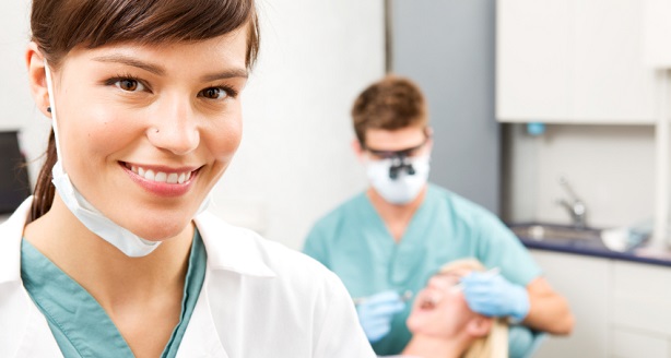 A portrait of a dental assistant smiling at the camera with the dentist working in the background